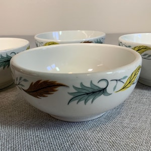 May include: Four white ceramic bowls with a leaf design in brown, teal, and yellow. The bowls have a rounded shape and are arranged on a gray surface. The leaf design is repeated around the rim of each bowl.