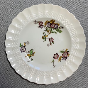 May include: White ceramic plate with a scalloped edge and a woven texture border. The plate features floral designs in shades of purple, green, and yellow. A decorative piece for serving or display.