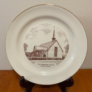 Könnte beinhalten: Ein weißer Zierteller mit goldenem Rand. Er zeigt eine braune Illustration einer Kirche mit Turm, Bäumen und Wolken. Der Text "First Methodist Church Fairbanks, Alaska" befindet sich darunter.