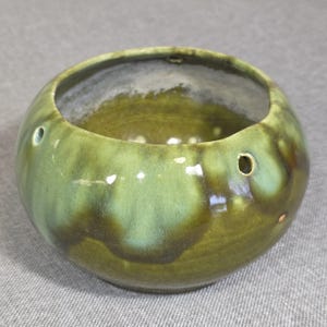 May include: A round ceramic bowl with a glossy, variegated glaze in shades of olive green, sage, and dark brown. The bowl features a wide, open top and two small, circular holes positioned symmetrically near the upper rim of the vessel.