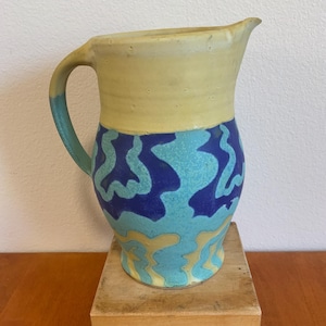 May include: A handmade ceramic pitcher with a yellow top and a turquoise handle. The body of the pitcher is decorated with blue and turquoise abstract patterns. The pitcher is sitting on a wooden block.