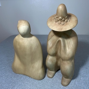 Könnte beinhalten: Zwei beige Keramikfiguren. Eine ist eine sitzende Figur mit abgerundeter Form. Die andere ist eine Figur, die einen Sombrero mit floralen Akzenten trägt. Die Figuren haben eine glatte, matte Oberfläche und werden auf einer grauen Oberfläche ausgestellt.