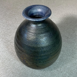May include: A round, bulbous vase with a narrow neck and a wide, flared rim. The surface features a dark, textured finish with deep blue and black tones, showing subtle horizontal ridges along the body and a metallic sheen on the rim.