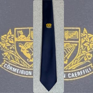 May include: A solid navy blue necktie featuring a small, centered gold and black embroidered crest. The crest depicts a harp and floral elements with the text CORMEIBION CAERFFILI visible in the background design.