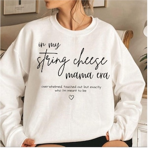 May include: White sweatshirt featuring the text "in my string cheese mama era" in black script. The text "overwhelmed touched out but exactly who I'm meant to be" and a heart symbol are below.