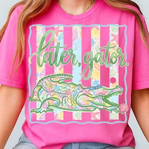 May include: A vibrant pink t-shirt with a colourful alligator graphic wearing sunglasses and holding a seashell. The words "later, gator" are written above the alligator in a playful font, set against a striped background.