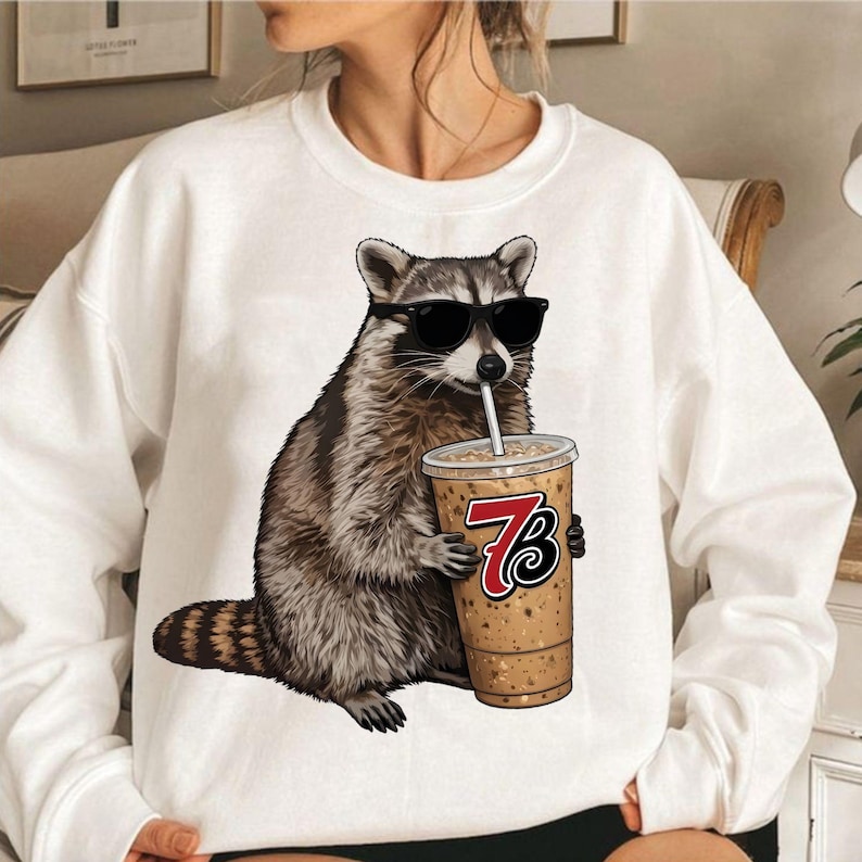 May include: White sweatshirt featuring a raccoon wearing sunglasses and holding a coffee drink with a straw. The coffee cup has the number 73 on it. The raccoon is brown, gray, and black.