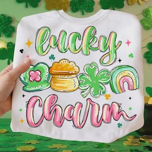 Puede incluir: Sudadera blanca con las palabras "Lucky Charm" en letras coloridas. El diseño incluye un trébol verde, una olla de oro, un arcoíris y otros gráficos temáticos del Día de San Patricio. La sudadera es de cuello redondo.