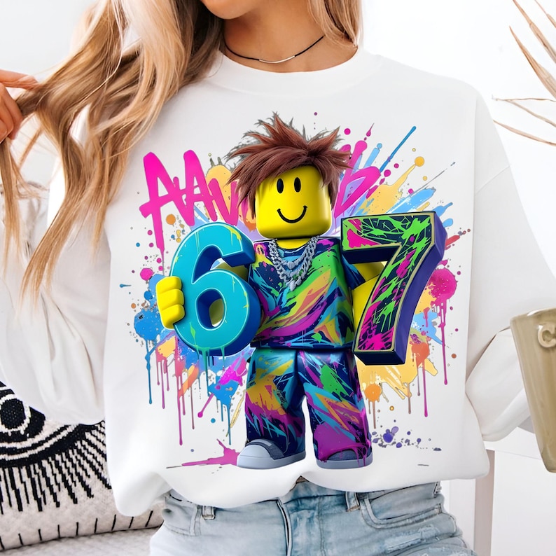 May include: White sweatshirt featuring a colorful graphic of a block figure holding the numbers 6 and 7. The figure has a yellow head with a smiley face and brown hair. The background has colorful paint splatters.