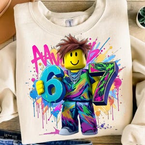 May include: Cream-colored sweatshirt featuring a colorful graphic of a block figure holding the numbers 6 and 7. The figure has brown hair and a yellow face. The graphic includes paint splatters in pink, blue, green, and yellow.