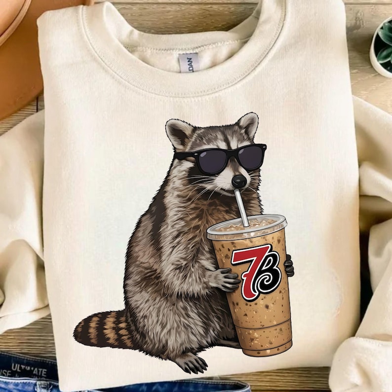 May include: Cream-colored sweatshirt featuring a raccoon wearing sunglasses and holding a coffee drink with a straw. The raccoon graphic is detailed with brown and gray fur. The coffee cup has a red and black logo.