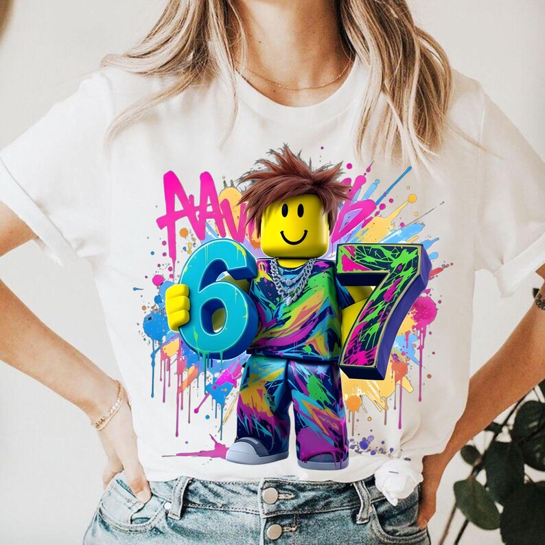 May include: White t-shirt featuring a colorful graphic of a block figure holding the numbers 6 and 7. The figure has a yellow head, brown hair, and a silver chain. The background has colorful paint splatters.
