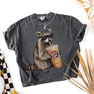 Puede incluir: Camiseta gris oscuro con un mapache con gafas de sol bebiendo de una taza de café. La taza tiene el número 73. La camiseta tiene las mangas enrolladas y cuello redondo.