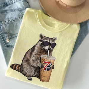 May include: A light yellow t-shirt featuring a raccoon wearing sunglasses and drinking from a coffee cup. The raccoon is brown and gray with a black nose and sunglasses. The coffee cup has a red and black logo.