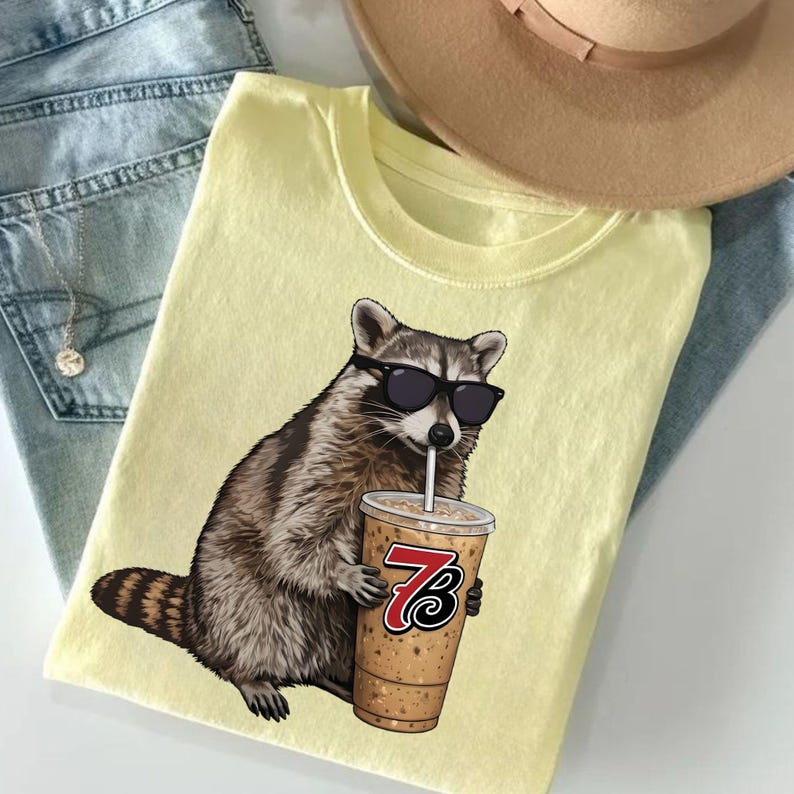 May include: A light yellow t-shirt featuring a raccoon wearing sunglasses and drinking from a coffee cup. The raccoon is brown and gray with a black nose and sunglasses. The coffee cup has a red and black logo.