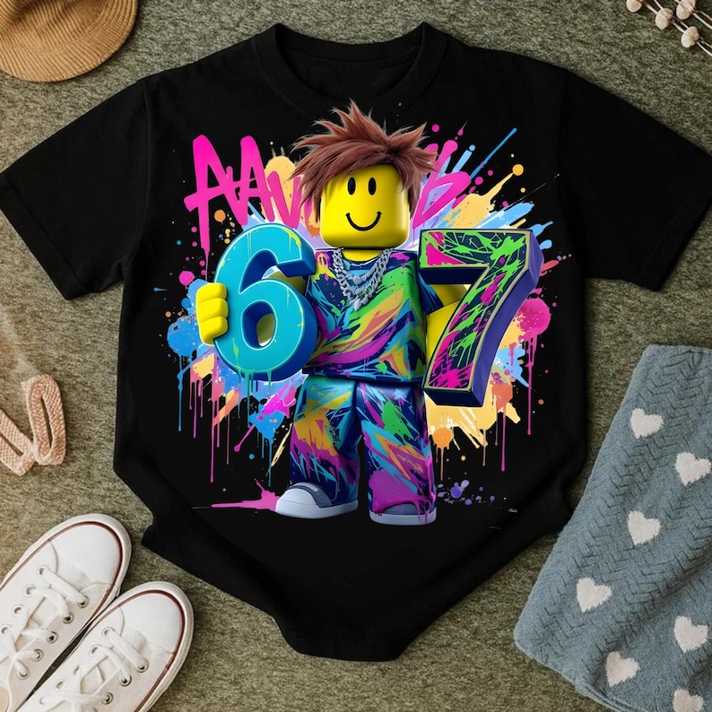 May include: Black t-shirt featuring a colorful graphic of a block figure holding the numbers 6 and 7. The figure has brown hair and a yellow head. The design includes neon paint splatters and the letters "AA".