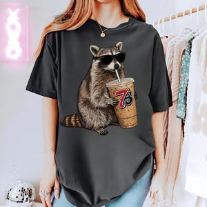 May include: Dark gray t-shirt featuring a raccoon wearing sunglasses and drinking from a coffee cup with the number 73 on it. The raccoon is brown and gray, and the coffee cup is tan with a white straw. The t-shirt is a relaxed fit.