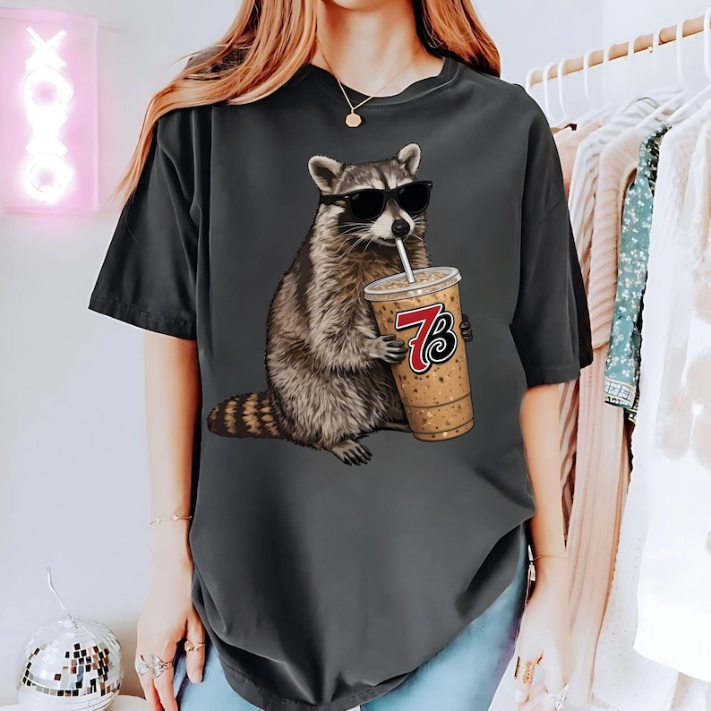 May include: Dark gray t-shirt featuring a raccoon wearing sunglasses and drinking from a coffee cup with the number 73 on it. The raccoon is brown and gray, and the coffee cup is tan with a white straw. The t-shirt is a relaxed fit.