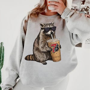 May include: A light gray sweatshirt featuring a raccoon wearing sunglasses and drinking from a beverage cup. The cup has the number 7 and the letter B printed on it. The raccoon is brown, gray, and white.