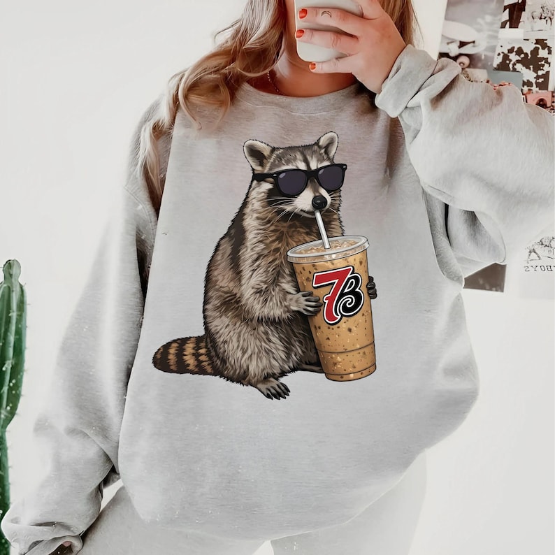 May include: A light gray sweatshirt featuring a raccoon wearing sunglasses and drinking from a beverage cup. The cup has the number 7 and the letter B printed on it. The raccoon is brown, gray, and white.