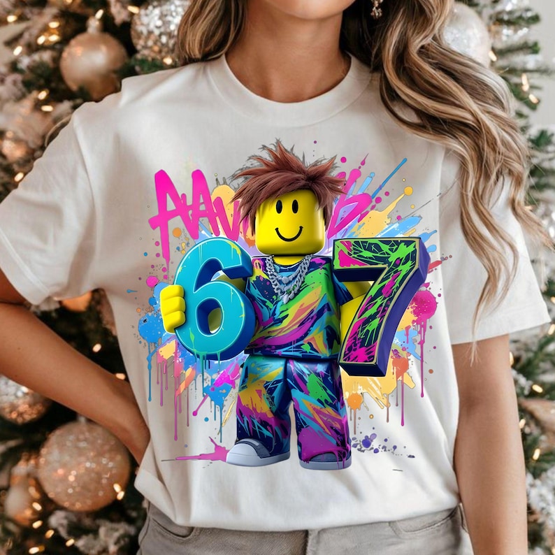 May include: White t-shirt featuring a colorful graphic of a Lego-style figure holding the numbers 6 and 7. The figure has a yellow head, brown hair, and a smiling face. The background has colorful paint splatters and the word "AAN".