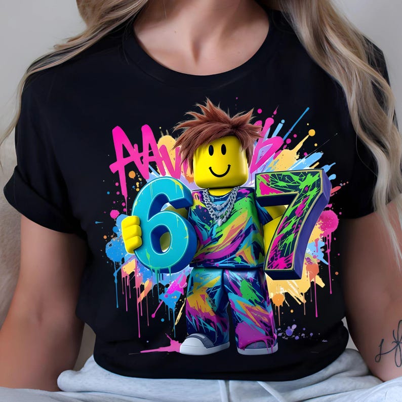 May include: Black t-shirt featuring a colorful graphic of a Roblox-style character holding the numbers 6 and 7. The character has a yellow head, brown hair, and a silver chain. The design includes neon paint splatters.