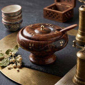 Può includere: Una ciotola per spezie in legno marrone scuro con coperchio abbinato e un piccolo cucchiaio di legno. La ciotola presenta una base a piedistallo e delicati motivi a intarsio color oro. Un piccolo pomello in ottone si trova al centro del coperchio e una tacca permette di riporre il cucchiaio.