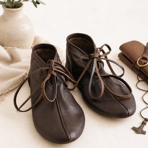 Könnte beinhalten: Ein Paar dunkelbraune Lederstiefeletten mit abgerundeter Zehenpartie, einer Mittelnaht auf der Vorderseite und dünnen Lederbändern, die oben gebunden werden. Die Stiefel haben eine weiche, flexible Konstruktion mit niedrigem Schaft und sichtbaren Nähten.