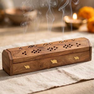 May include: A rectangular wooden incense burner box with a hinged lid featuring intricate laser-cut geometric patterns. The front side is decorated with three small, inlaid brass elephant figures. Wisps of white smoke rise from the top.
