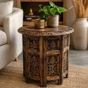 May include: A round wooden side table featuring intricate hand-carved floral patterns on the top edge and side panels. The table has a dark brown finish and a multi-sided base. A gold-toned metal candle holder and a potted green plant sit on top.