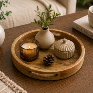 May include: A round wooden serving tray with cutout handles containing a small amber glass candle holder with a lit candle, a beige ceramic bud vase with green leaves, a small lidded beige ceramic jar, and a single brown pinecone.