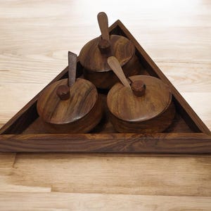 May include: A wooden spice rack set with three small, round lidded bowls and spoons. The bowls are set in a triangular tray, all made of dark brown wood. The spoons are inserted into the lids. The set is designed for kitchen use.
