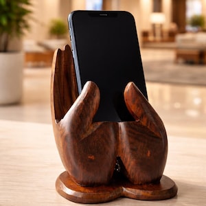 Puede incluir: Un soporte para teléfono de madera marrón oscuro tallado con la forma de dos manos ahuecadas que descansan sobre una base plana. Las manos están posicionadas para sostener un teléfono inteligente negro en posición vertical.