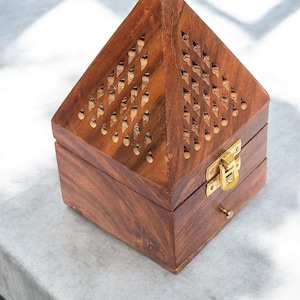 Peut inclure: Boîte en bois en forme de pyramide avec un couvercle à charnière et un motif décoratif de petits trous. La boîte est en bois brun foncé et possède un loquet doré et un petit tiroir. La boîte est posée sur une surface gris clair.