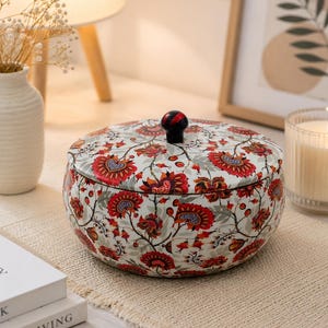 May include: A round metal storage tin with a lid featuring a vibrant floral pattern in shades of red, orange, and grey on a white background. The container has a small, dark, rounded knob on top of the lid for easy opening and closing.
