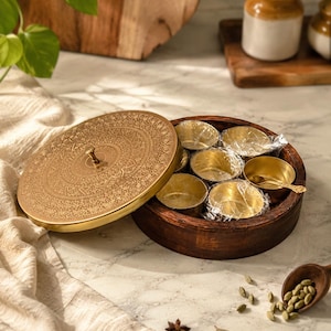 Può includere: Una scatola per spezie rotonda in legno con un coperchio in ottone rimovibile caratterizzato da intricati motivi incisi. L'interno contiene diverse piccole ciotole in ottone, alcune avvolte in plastica trasparente, e un cucchiaino in ottone.