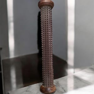 Puede incluir: Un rodillo de pie de madera marrón con un diseño cilíndrico con púas. El rodillo tiene una parte superior e inferior redondeadas y está diseñado para masajes. El rodillo mide aproximadamente 30 cm de alto.