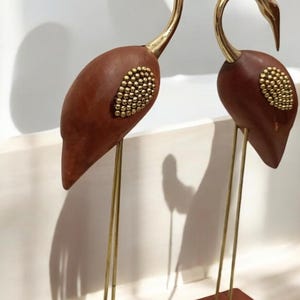 Puede incluir: Dos esculturas decorativas de grullas. Las grullas tienen cuerpos marrones con detalles dorados y patas de latón. Los cuerpos están adornados con pequeños adornos dorados. Las esculturas se alzan sobre bases cuadradas marrones.