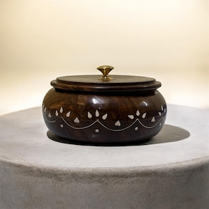 Puede incluir: Una caja de madera marrón oscuro con tapa y pomo de latón. La caja está decorada con un patrón blanco en forma de corazón alrededor de la base. La caja está sobre una superficie gris.