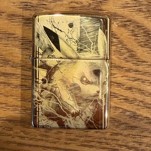May include: A gold-colored metal lighter featuring a detailed illustration of Pikachu from the Pokémon franchise. The design is etched into the metal, showcasing the character's face and lightning bolt details. The lighter is rectangular with a hinged lid.