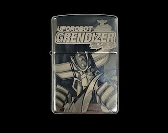 Puede incluir: Un encendedor de metal rectangular con acabado pulido que presenta un grabado del personaje Uforobot Grendizer. El texto UFOROBOT GRENDIZER es claramente visible en la parte superior, junto a caracteres japoneses.