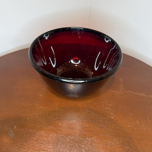 Può includere: Una ciotola di vetro rosso rubino dalla forma arrotondata. La ciotola ha un bordo scuro e una finitura liscia e lucida. L'interno della ciotola è di un rosso intenso. La ciotola è su una superficie di legno.
