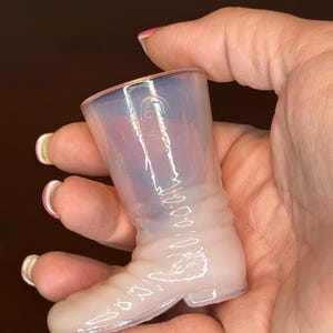 May include: A small, translucent glass boot with a light pink and blue hue. The boot has a detailed design, including a heel, sole, and stitching details. The glass has a glossy finish.