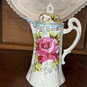 Puede incluir: Una cafetera de porcelana blanca con una gran rosa rosa pintada en el frente. La jarra tiene un borde azul claro y detalles dorados. El asa es curva y la tapa tiene un pequeño adorno decorativo. La jarra mide aproximadamente 20 cm de alto.