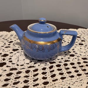 May include: A small, blue ceramic teapot with a gold band of decorative detailing. The teapot has a matching lid with a small knob and a spout. The handle is rectangular. The teapot is sitting on a white, crocheted doily.