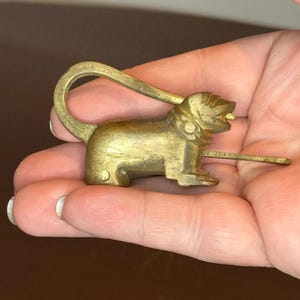 Puede incluir: Un candado vintage de latón con forma de león. El candado dorado presenta un asa curvada y una cerradura. El león está representado en posición sentada con la boca abierta. El candado parece ser de metal.
