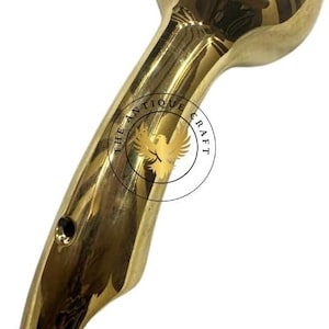 May include: A polished brass object with a rounded head and curved handle. The handle features a circular logo with an eagle and the text "THE ANTIQUE CRAFT". The object has a smooth, reflective surface.