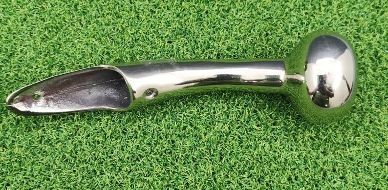 May include: A polished, silver-colored metal tool with a rounded end and a scoop-shaped opening. The tool has a curved handle and is designed for a specific purpose. The object is set against a green background.