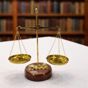 May include: A brass balance scale with two round pans suspended by chains. The scale sits on a dark wooden base with gold star accents. The background features a blurred bookshelf, suggesting a legal or academic theme.