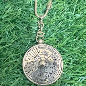 Op de afbeelding: Een messingkleurige eeuwige kalender sleutelhanger. De cirkelvormige kalender heeft de tekst "PLACE YEAR OVER MONTH" en "TALIC MONTH FOR LEAP YRS". De sleutelhanger zit aan een ketting met een sluiting.